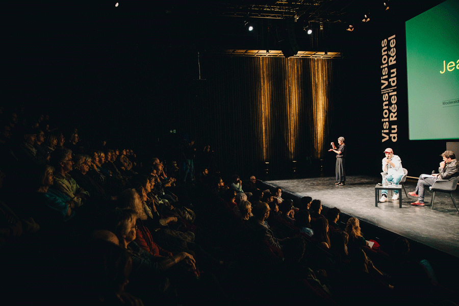 Un intérieur de théâtre. La salle est pleine obscure. A droite, sur la scène éclairée, trois personnages. Au centre, deux hommes assis sur des chaises. Le premier homme regarde le deuxième homme qui tient un micro et a le visage tourné vers le public. Le deuxième homme est habillé en couleurs claires et porte une casquette rouge. A leur droite, une femme vêtue de noir se tient debout. Elle est éclairée, regarde la public et bouge ses deux mains à hauteur du visage. Sur le mur du fond, on distingue une partie d’un écran vert et une grand inscription verticale « Visions du réel / Visions du réel » blanche sur fond noir.