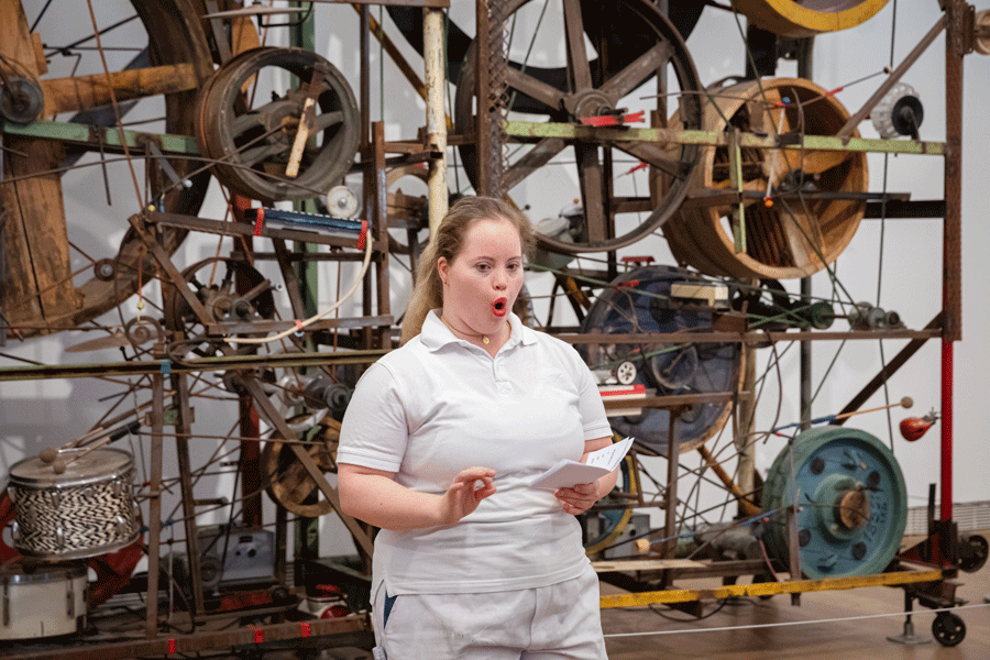 Im Vordergrund eine Schauspielerin des machTheaters in weisser Hose und weissem Shirt. Sie ist am Sprechen. Hinter ihr ist eine grosse Klangmischmaschine von Jean Tinguely, die Méta-Harmonie 2. 