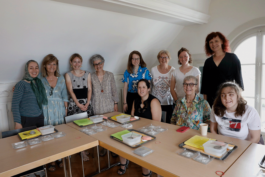 Dans un espace intérieur, 11 femmes de différents âges se tiennent autour de deux tables. Trois d’entre elles sont assises. Toutes regardent l’objectif et sourient. Sur les tables, il y a du matériel, entre autres des plateaux porte-assiettes, des récipients ronds avec un liquide clair et une passoire à thé, des récipients carrés en verre avec une matière blanche, des éponges couleur saumon, ainsi que des carrés de tissu épais jaune et vert clair.