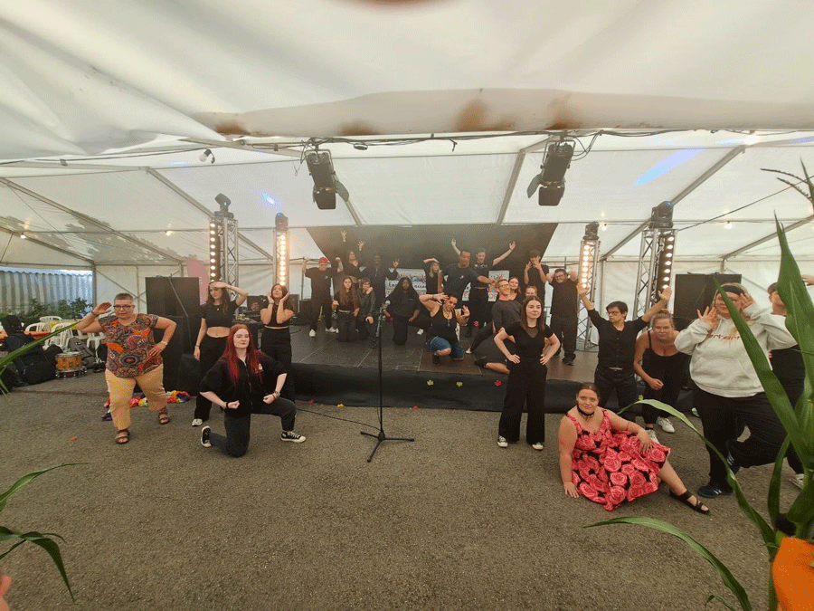 Une scène dans une tente de fête. Une vingtaine de jeunes personnes sont debout, accroupies ou assises sur scène et sur le sol. Elles regardent devant elles en riant. Quelques personnes ont les mains sur les hanches ou lèvent les bras. 