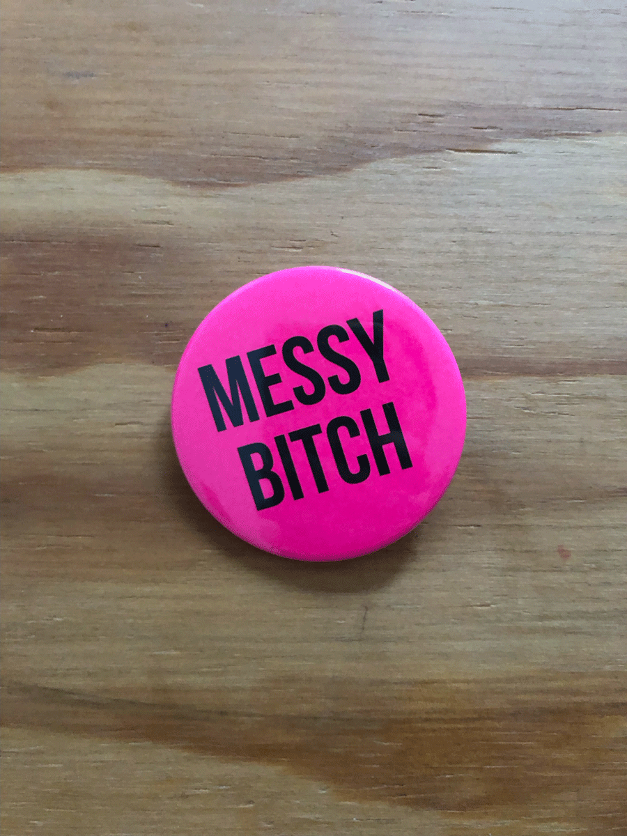 Das Bild zeigt einen pinken Button. Der Button ist rund und hat etwa einen Durchmesser von sieben Zentimetern. Er trägt eine schwarze Aufschrift, die sagt: Messy Bitch (deutsch: chaotische Schlampe).