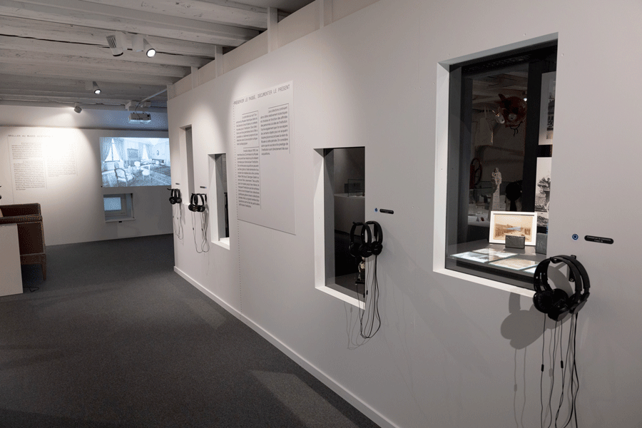 Un mur intérieur blanc avec quatre vitrines encastrées à la manière de fenêtres, flanquées de casques d’écoute fixés au mur. Dans la vitrine au premier plan, cinq images gravées. Au milieu du mur, un texte de salle noir sur fond blanc. 