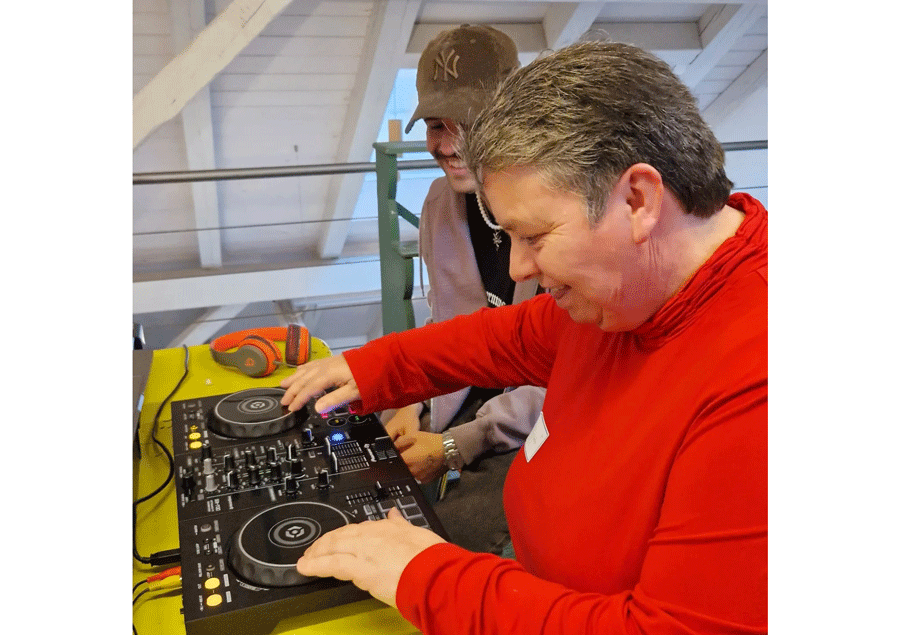 Eine Frau mit kurzen grauen Haaren und in rotem Pullover berührt mit den Finderspitzen die beiden Stellen an einem Mischpult, die beim Scratchen bewegt werden. Sie und der junge Mann mit Baseballkappe neben ihr blicken konzentriert darauf und lächeln.