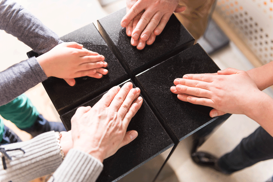 Vier Handpaare von oben gesehen, jeweils eine Handfläche liegt auf der anderen. Die Hände ruhen auf einem schwarzen Sockel, die Finger sind zur Mitte hin gerichtet. Der Sockel ist in vier Quadrate unterteilt, die sich nicht berühren. 