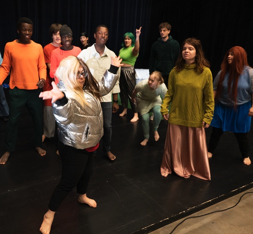 Sur fond sombre d’une scène à même le sol, 11 jeunes personnes vêtues de couleurs et pieds nus prennent différentes poses. Au premier plan à gauche, une jeune femme blonde avec une veste gris métallique lève les bras en forme de V. 